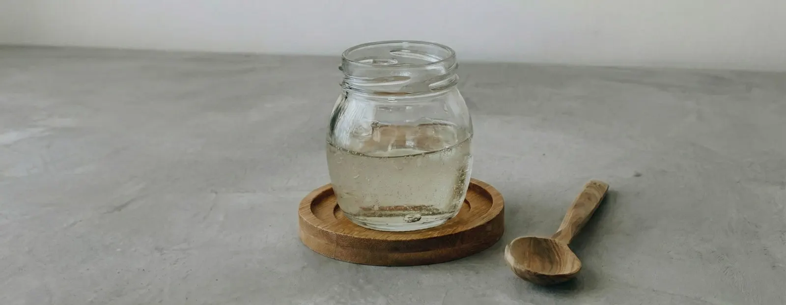 A glass jar of clear, honey-like liquid on a wooden coaster beside a wooden spoon