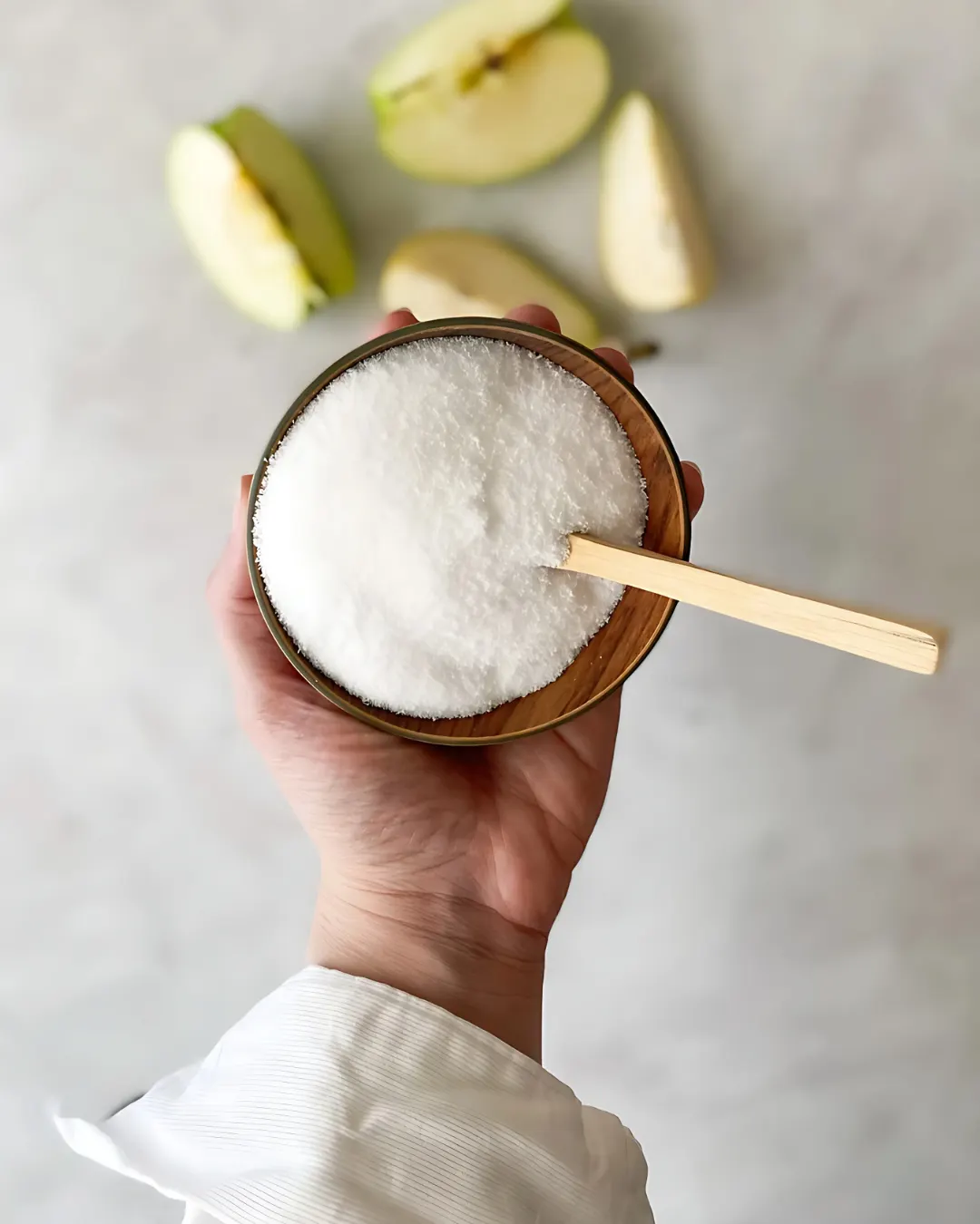 A hand holding a small bowl of white sweetener powder above sliced apples