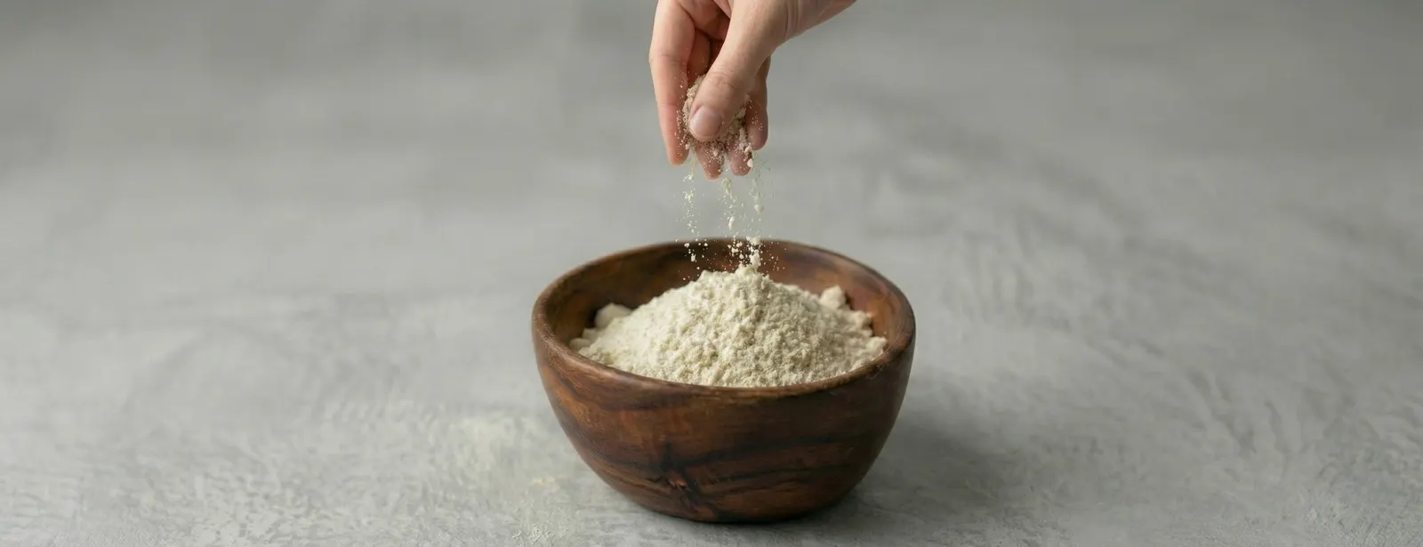 A hand sprinkling pale powder into a wooden bowl on a grey surface