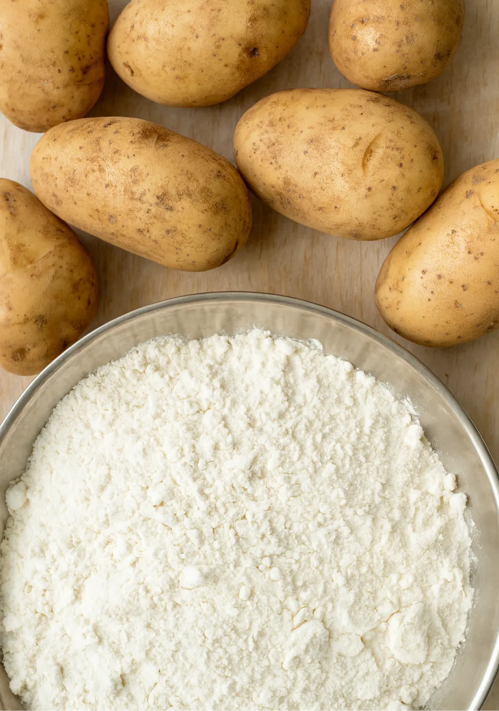 Whole potatoes beside a bowl of white starch powder on a wooden surface