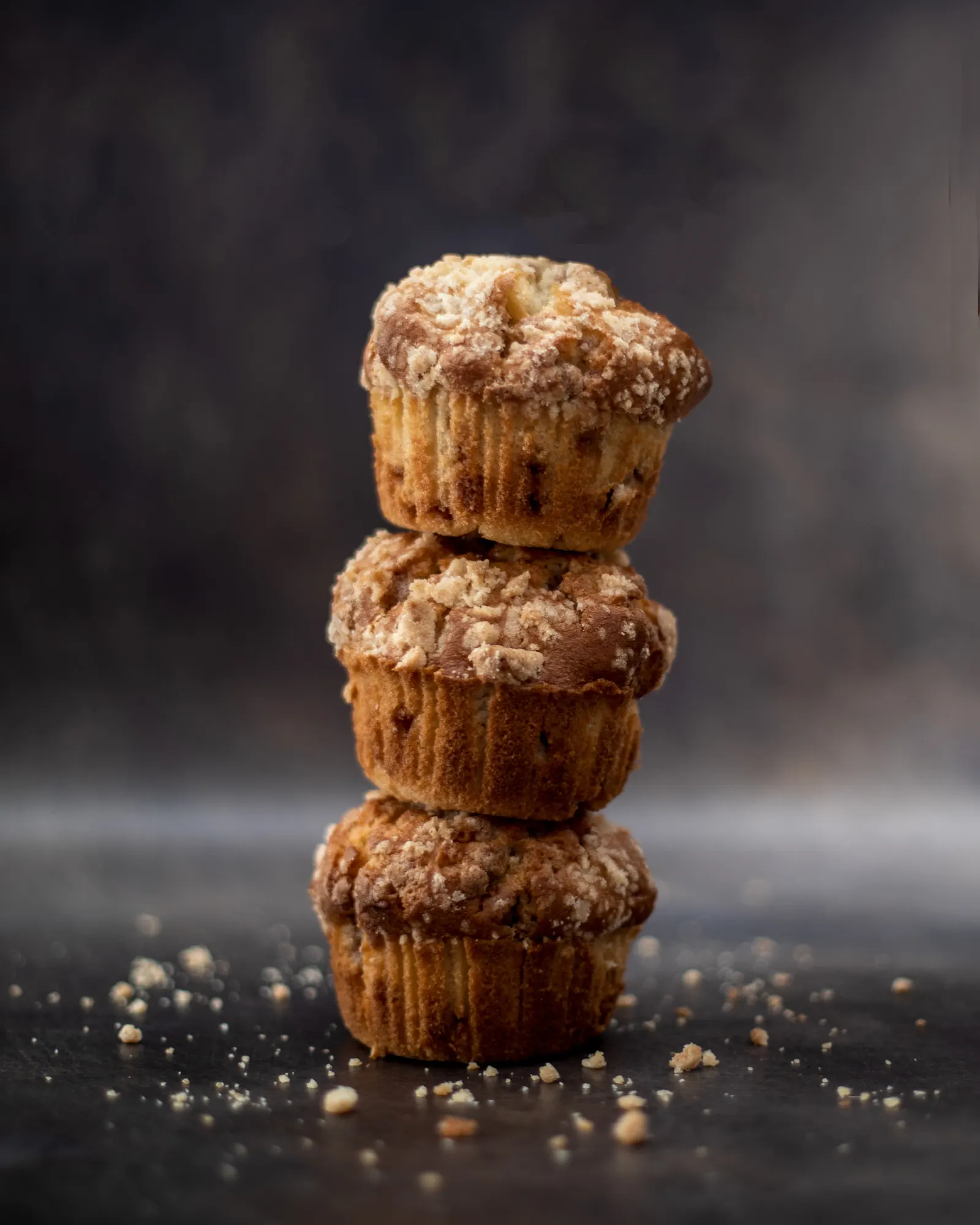 Three crumb-topped muffins stacked on a dark surface